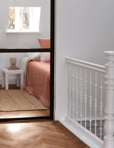Overloop met visgraatparketvloer en witte klassieke trapleuning, met doorkijk via zwarte stalen deuren naar een slaapkamer met terracotta beddengoed en jute vloerkleed