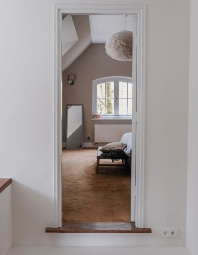 Overloop met houten trapleuning en doorkijk naar sfeervolle slaapkamer met veren hanglamp en taupe muren