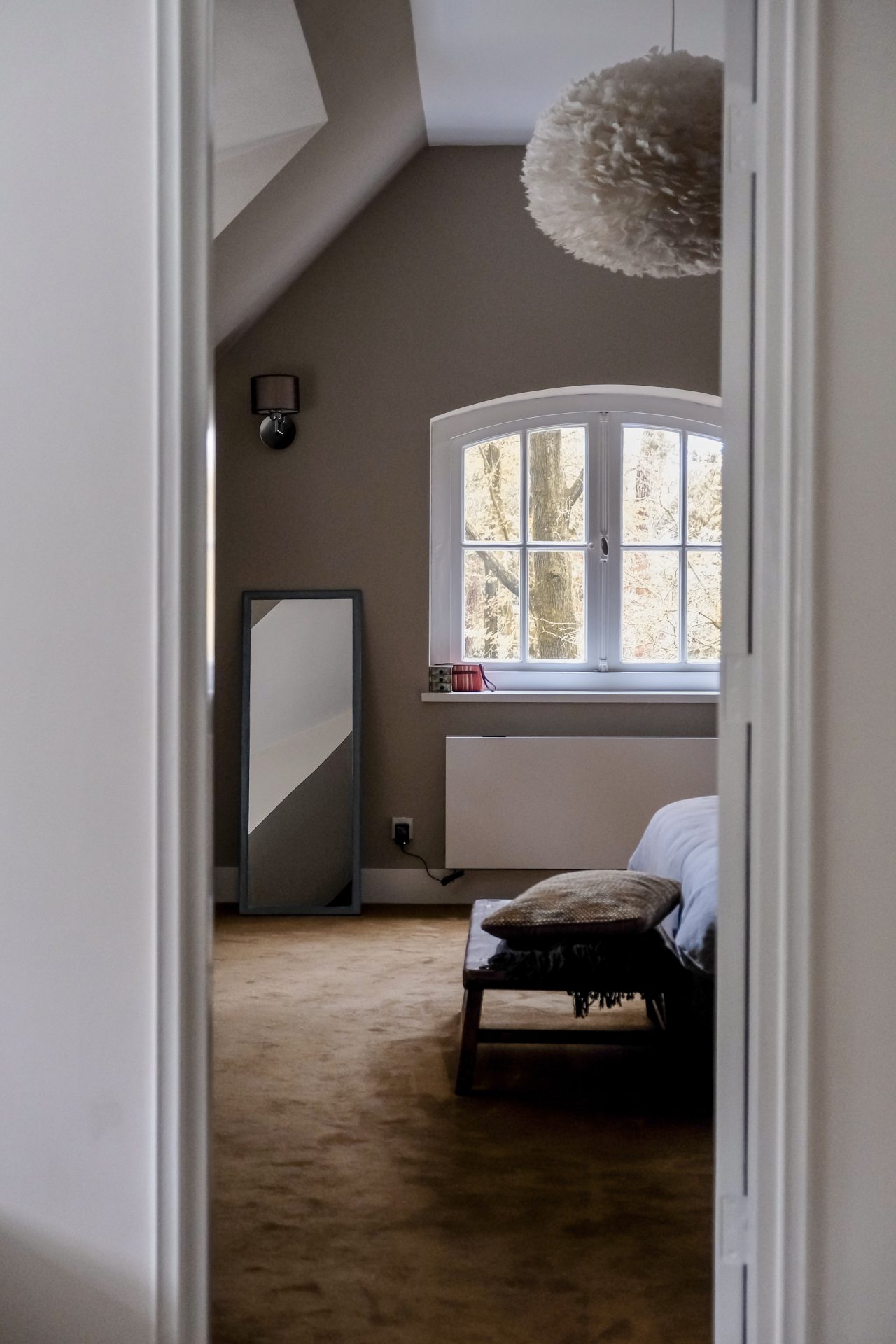 Slaapkamer op zolder met rondboogvenster, taupe muren, staande spiegel en sfeervolle veren hanglamp in Hilversum