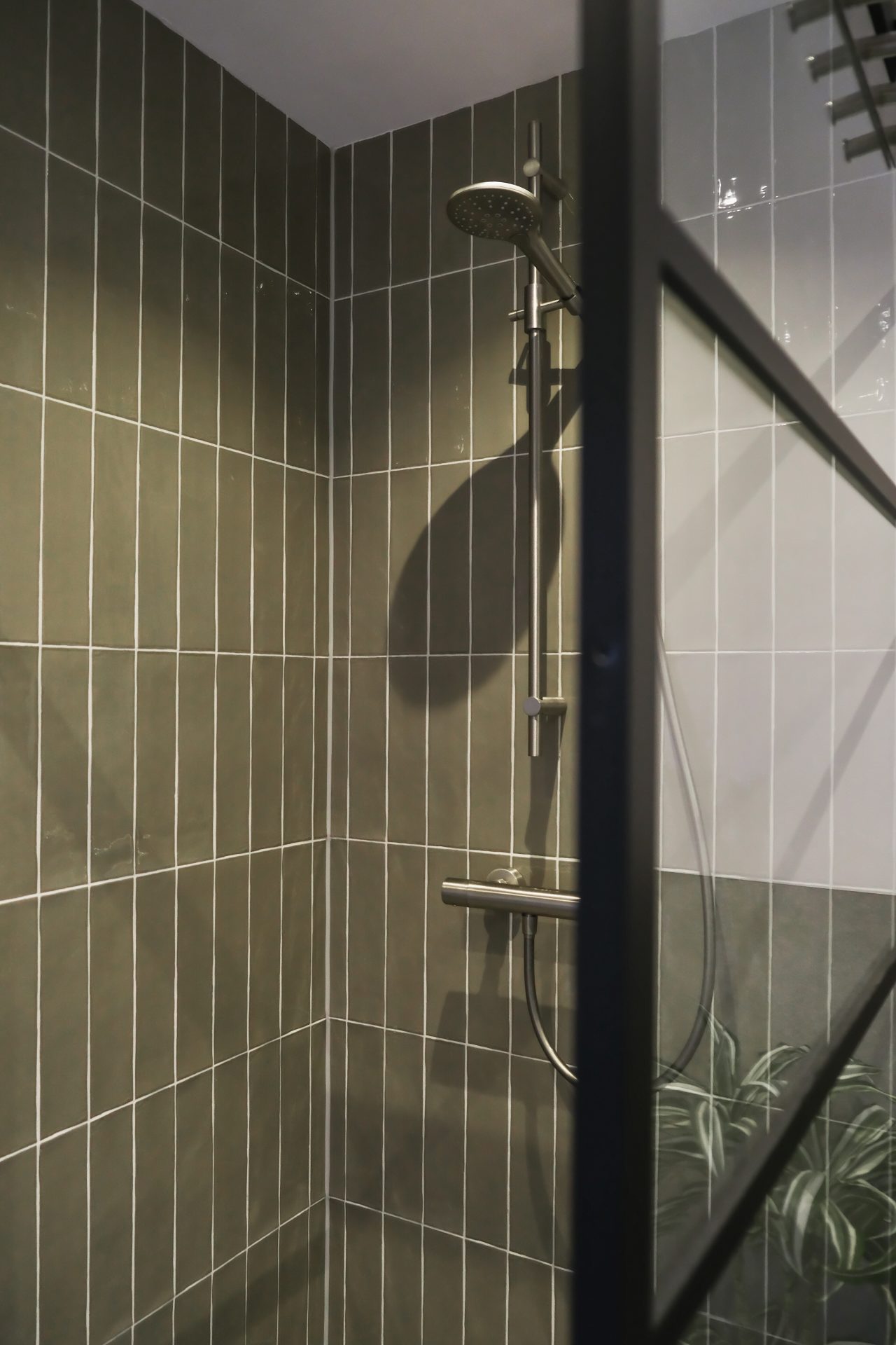 Detailopname van inloopdouche met olijfgroene smalle tegels, stalen doucheset en zwart stalen glaswand