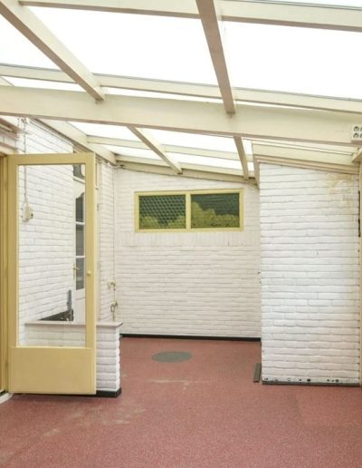 Verouderde glazen serre met wit geschilderde bakstenen muren, rode rubbervloer, gele houten deuren en een doorkijk naar de woonkamer