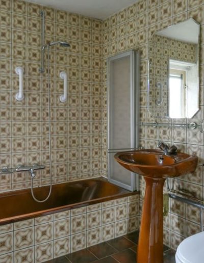 Sterk verouderde badkamer voor renovatie met van vloer tot plafond bruinbeige patroontegels uit de jaren 70, bruine ligbad met douchecombinatie, bruine zuil-wastafel en toilet