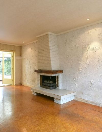 Ruime woonkamer met terracotta kurkvloer, witte bakstenen schouw in hoek, grove spuitpleisterwand, open keuken met bruine kastjes en doorgangsdeur naar de serre