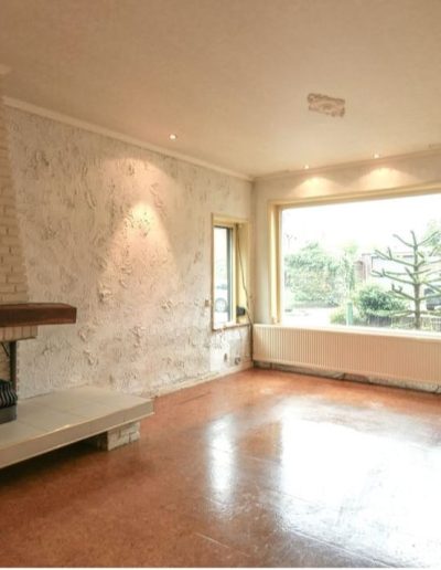 Lege woonkamer met terracotta kurkvloer, wit geschilderde bakstenen schouw met houten bovendorpel, grof gepleisterde muren en een groot raam aan de straatkant