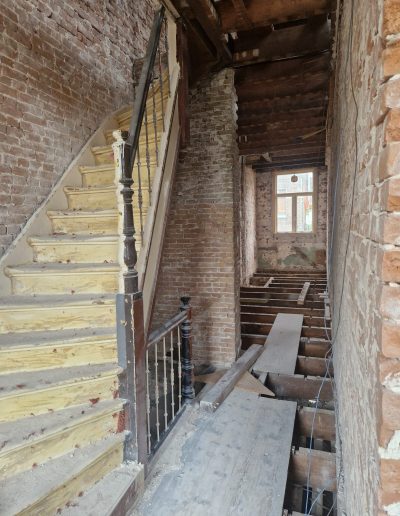 Originele geel geschilderde houten trap met donkere gedraaide balusters in een gestript trappenhuis met blootgelegde bakstenen muren, blootgelegde vloerbalken en blauwe elektrakabels