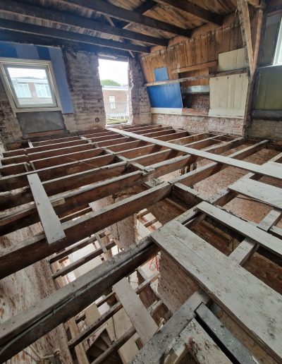 Interieur van een gestripte bovenverdieping van een oud pand met blootgelegde houten vloerbalken, zichtbare bakstenen muren en een houten dakconstructie, met doorkijk naar buiten via twee ramen