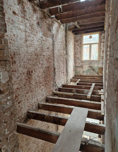 Lange smalle gang van een pand in renovatie met volledig blootgelegde bakstenen muren, houten vloerbalken zonder vloerafwerking en een raam aan het einde dat zorgt voor daglicht