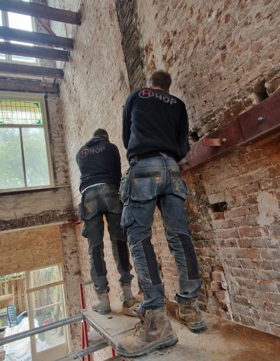 Twee medewerkers van Bouwbedrijf Hop staan op een steiger en verwerken een rode stalen HEA-balk in een opening in een bakstenen muur, met een glas-in-loodraam op de achtergrond
