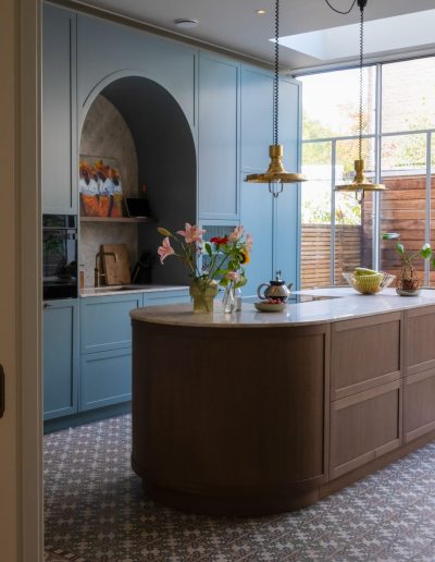 Ruime keuken met blauw-groene shaker kastjes, een gewelfd nis, een ovaal houten kookeiland met marmeren blad, gouden hanglamp en patterned tegelvloer