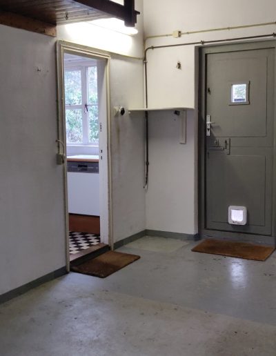 Situatie voor renovatie: ruimte met betonvloer, houten dakconstructie, groene buitendeur met kattenluik en open deur naar bijkeuken